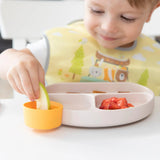 A child in a bib smiles at fun shapes and colors on a divided plate with apple slices, strawberries, and the Bumkins Tutti-Frutti Dippers.