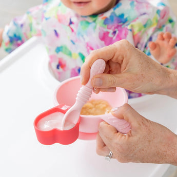 An adult uses Bumkins Silicone Little Dipper in Tutti-Frutti to feed a baby in a high chair, adding fun with the colorful bib.