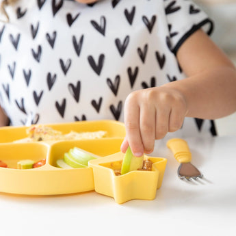 A child in a heart shirt enjoys apples with sauce from a Bumkins star-shaped bowl on a yellow tray, surrounded by fun colors.