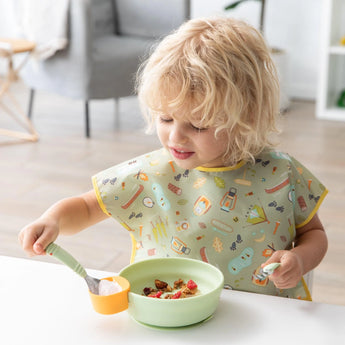 A child in a Bumkins Tutti-Frutti bib enjoys a meal with the Silicone Little Dipper in the bright dining room.