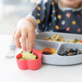 A child joyfully dips a cucumber slice in a pink, food-grade cup from Bumkins Silicone Little Dipper 3-Pack: Tutti-Frutti, beside fun treats.