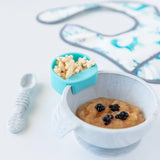 Bumkins Silicone Little Dipper Round in Gumdrop and safety-tested dishware, spoon, bib, plus a cereal container on white surface.