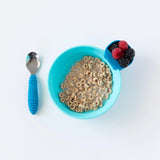 A blue cereal bowl with Bumkins Silicone Little Dipper holders for berries sits next to a spoon with a blue handle on a white background.