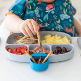 Child dipping pretzels into chocolate on a Gumdrop bib, with eggs and fruit on safety-tested Bumkins Silicone Little Dipper dishware.