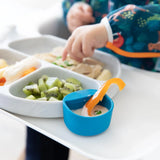 A toddler in a colorful bib sits at a white highchair, picking up food from a divided plate with sliced kiwi and chicken. In the foreground, showcasing eco-friendly dip holders, is the Bumkins Silicone Little Dipper Round in blue with dip and a carrot stick.