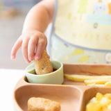 A childs hand dips a nugget into sauce on a Bumkins Silicone Little Dipper Gumdrop plate with fries and nuggets.