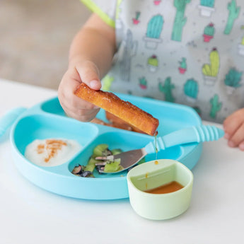 A child uses a Bumkins Silicone Little Dipper to enjoy breadsticks with sauce, yogurt, sliced veggies, and a trusty fork.