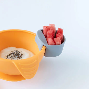 Yellow bowl with yogurt and chia seeds, plus a Bumkins gray Silicone Little Dipper holding watermelon on a white background.