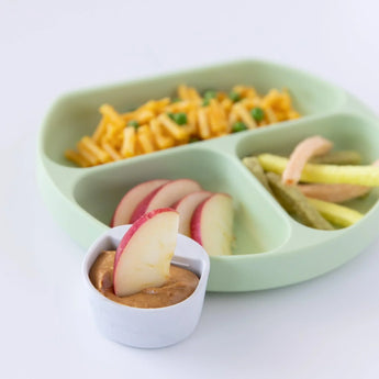 A green divided plate holds pasta, peas, veggie sticks, with a Bumkins Silicone Little Dipper filled with apple slices and PB.