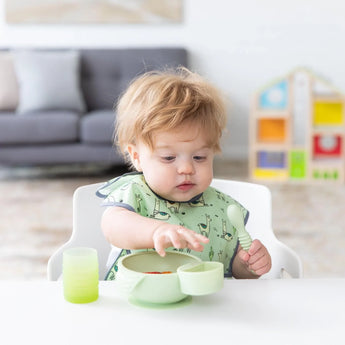 A curly-haired toddler holds a spoon near Bumkins Silicone Little Dipper Round 3-Pack: Taffy, with a couch and toy shelf in the background.