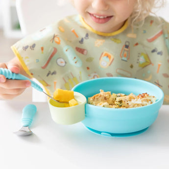 Child with a colorful bib uses Bumkins Silicone Little Dipper in Taffy, featuring a detachable snack cup for fruits.