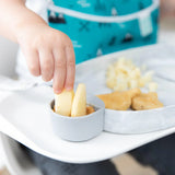 A child dips apple slices into peanut butter using Bumkins Silicone Little Dipper Round 3-Pack: Taffy, with snacks in the background.