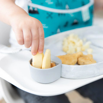 A child dips apple slices into peanut butter using Bumkins Silicone Little Dipper Round 3-Pack: Taffy, with snacks in the background.