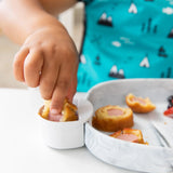 Child in blue mountain-patterned shirt dips mini corn dog into sauce in a Bumkins Silicone Little Dipper, bites scattered on plate.