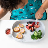 A child enjoys corn dog pieces, fruits, and broccoli with ketchup from Bumkins Silicone Little Dipper, wearing a blue mountain bib.