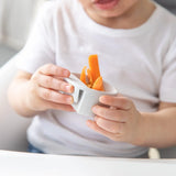 A child in a white shirt holds a cup with carrot sticks, sitting at a table with Bumkins Silicone Little Dipper Round 3-Pack: Taffy.