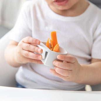 A child in a white shirt holds a cup with carrot sticks, sitting at a table with Bumkins Silicone Little Dipper Round 3-Pack: Taffy.