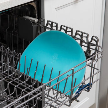 Open dishwasher with blue plate in the lower rack, next to a Bumkins Silicone Sensory Placemat: Blue. Kitchen cabinets visible behind.