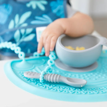 A baby reaches for pasta on Bumkins blue silicone sensory placemat, with a gray spoon attached to a teething toy.