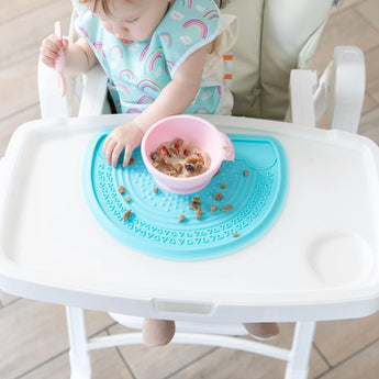 A baby uses a pink spoon and bowl on a Bumkins Silicone Sensory Placemat in blue, designed to gather food pieces.