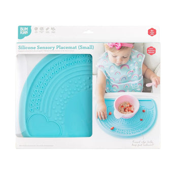 A baby happily uses a Bumkins Silicone Sensory Placemat: Blue, with the nonslip base keeping it secure, featuring a cheerful rainbow design.