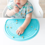 Baby eagerly eats finger food on a blue Bumkins Silicone Sensory Placemat, wearing a dinosaur-patterned bib in a high chair.