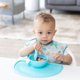 A toddler in a bib happily eats cereal from a blue bowl on a Bumkins Silicone Sensory Placemat.