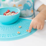 A childs hand explores cereal on a Bumkins Silicone Sensory Placemat: Blue, with a dinosaur bib and cereal bowl nearby.