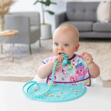 A baby chews a blue teething toy on a Bumkins Silicone Sensory Placemat: Blue, snacks scattered, in a cozy living room.