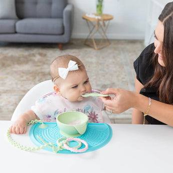 A woman feeds a baby wearing a headband and bib, with food on a Bumkins Silicone Sensory Placemat: Blue in front of them.