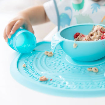 Child in a dinosaur shirt spills a blue cup on a cereal-filled Bumkins Silicone Sensory Placemat: Blue.