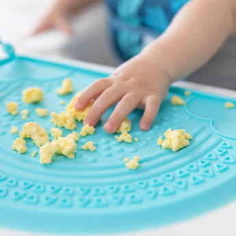 A child plays with scrambled eggs on a bright blue Silicone Sensory Placemat from Bumkins.