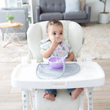 Baby in a high chair, using a purple bowl and spoon on a Bumkins Silicone Sensory Placemat: Gray, wearing a floral bib.