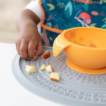 A child wearing a colorful bib is seated at a table, picking up apple pieces from the Bumkins Silicone Sensory Placemat: Gray, with an orange bowl nearby. The setting is bright and focused on eating.