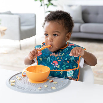 A toddler eats from an orange bowl on a Bumkins Silicone Sensory Placemat: Gray in a bright living room.