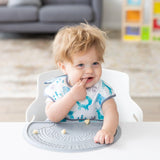 A toddler with messy hair sits in a high chair, wearing a bib, exploring food on a Bumkins Silicone Sensory Placemat: Gray.