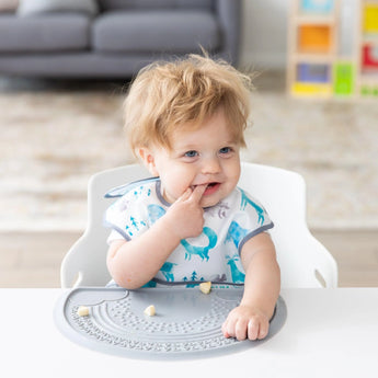 A toddler with messy hair sits in a high chair, wearing a bib, exploring food on a Bumkins Silicone Sensory Placemat: Gray.