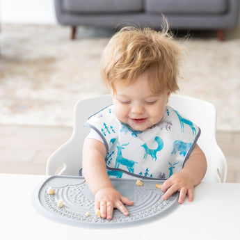 A curly-haired baby in a patterned bib plays with food on a Bumkins Silicone Sensory Placemat: Gray in their high chair.