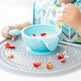 A child in a patterned bib spills strawberries and yogurt from a blue bowl onto a textured tray with the Bumkins Gray Silicone Sensory Placemat.