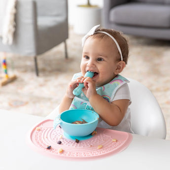 A baby with a headband smiles in a high chair, biting a spoon. A bowl rests on the Bumkins Silicone Sensory Placemat: Pink.