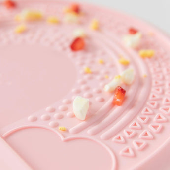 Close-up of a textured pink baby plate with crumbs and food bits on Bumkins Silicone Sensory Placemat: Pink.