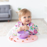 A toddler with a colorful bib smiles while holding food on a Bumkins Silicone Sensory Placemat: Pink, with a purple bowl and snacks.