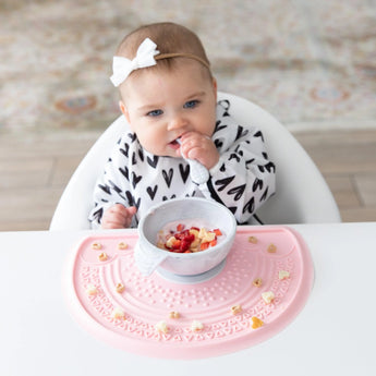 A baby, in a heart-patterned outfit with a white bow, joyfully engages in sensory play while seated in a high chair. She holds a spoon and eats chopped fruit from an eco-friendly Silicone Sensory Placemat: Pink by Bumkins on a patterned carpet backdrop.