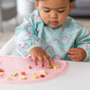 A toddler in a rainbow bib explores food on a pink highchair, enhanced by the Bumkins Silicone Sensory Placemat: Pink.