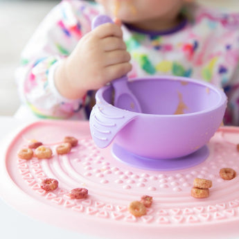 A child enjoys sensory play with cereal on a Bumkins Silicone Sensory Placemat: Pink, holding a spoon with a purple bowl.