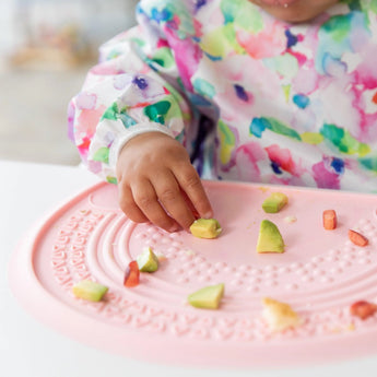 A child in a colorful bib reaches for avocado on a vibrant pink Bumkins Silicone Sensory Placemat.