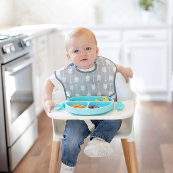 A baby in a high chair wears a Bumkins SuperBib®: Arrow, enjoying snacks from a blue plate in a vibrant, machine-washable kitchen.