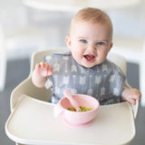 A baby in a high chair, wearing a Bumkins SuperBib®: Arrow, enjoys their meal with a pink bowl and spoon.