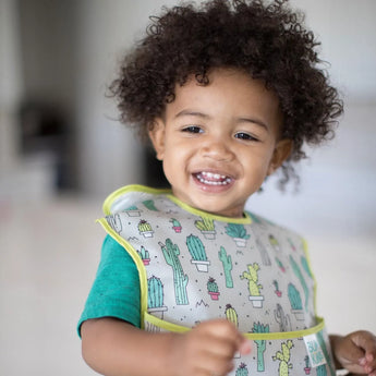 Curly-haired toddler wears a Bumkins SuperBib®: Cacti, standing inside.