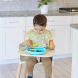 A toddler in a high chair munches from a blue plate, wearing a Bumkins SuperBib®: Camp Gear. The kitchen has white cabinets and a plant.